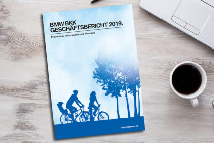 Der BMW BKK Geschäftsbericht 2019 liegt neben einer Tasse Kaffee, Handy und PC auf einem Tisch.
